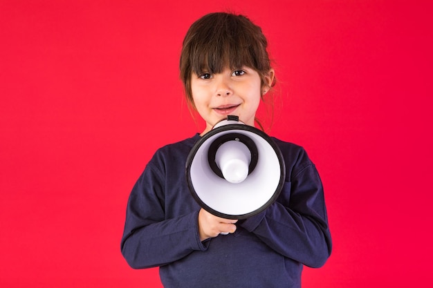 Blackhaired girl in a blue sweater and white pants speaking through a white megaphone on a red background