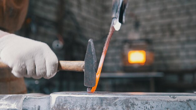 The blacksmith bends iron rod on the anvil with hammer, metal work, close up, telephoto