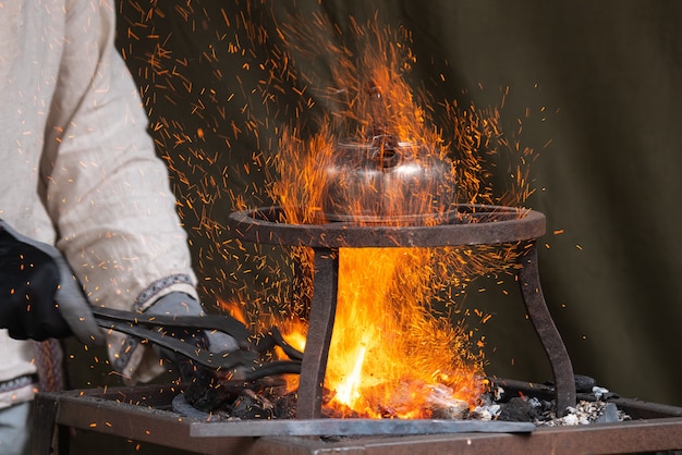 Blacksmith heating up metal in red hot coals while tea pot boiling water on top.