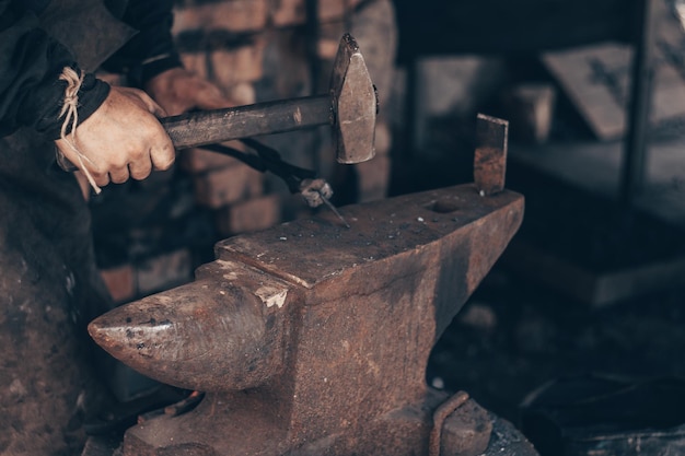 Foto fabbro che lavora il metallo con martello e tenaglie sull'incudine nella fucina maniscalco che colpisce il ferro in officina lavorazione dei metalli