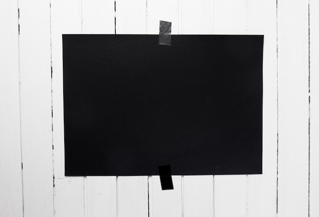 Blank black poster hanging on a tape on white wooden plank wall. Template background for your design.