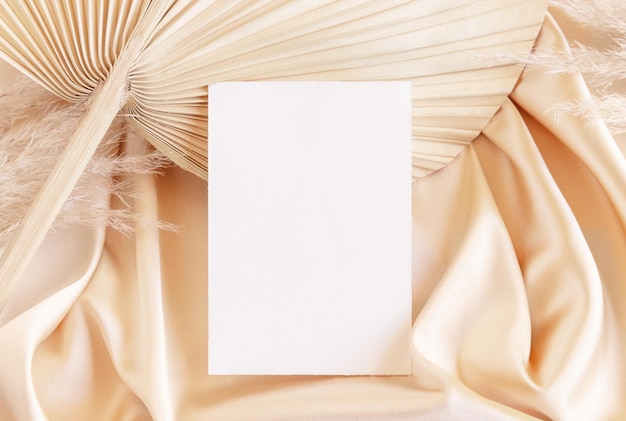 Blank card on beige satin fabric and dried palm leaf close up greeting or wedding mockup