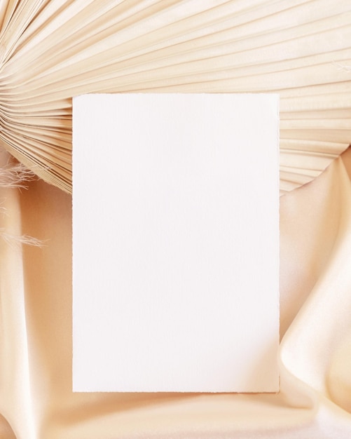 Blank card on beige satin fabric and dried palm leaf close up greeting or wedding mockup