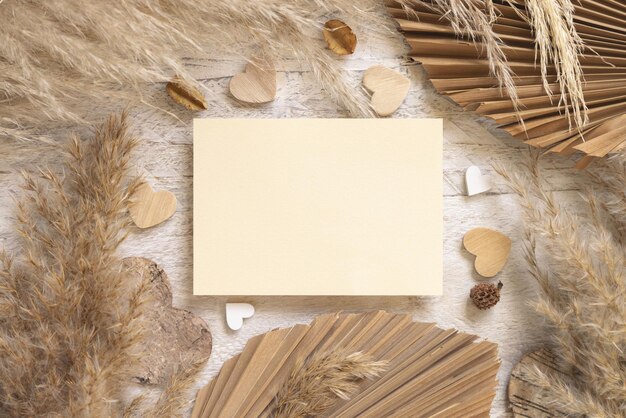 Blank card on white wooden table near dried pampas grass and hearts top view. Boho scene with horizontal paper card, flat lay. Bohemian Valentines mockup