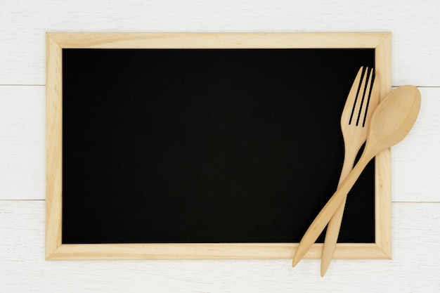 白い木の板の背景に木のスプーンとフォークと空白の黒板