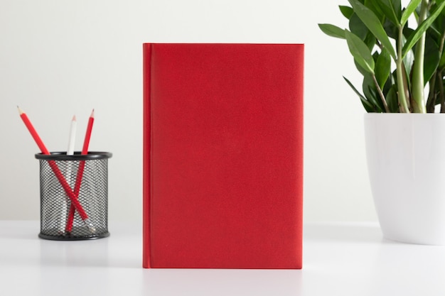 Foto copertina vuota di un libro rosso o di un diario su sfondo bianco con matite e una pianta su un vaso