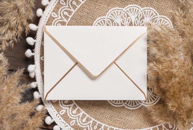 Blank envelope on bohemian jute placemat near pampas grass top view. Boho rustic scene with greeting or Invitation envelope mockup, flat lay.