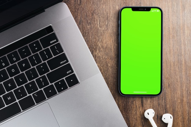 Blank green screen smartphone on wooden background with a computer beside it. Chroma Key.