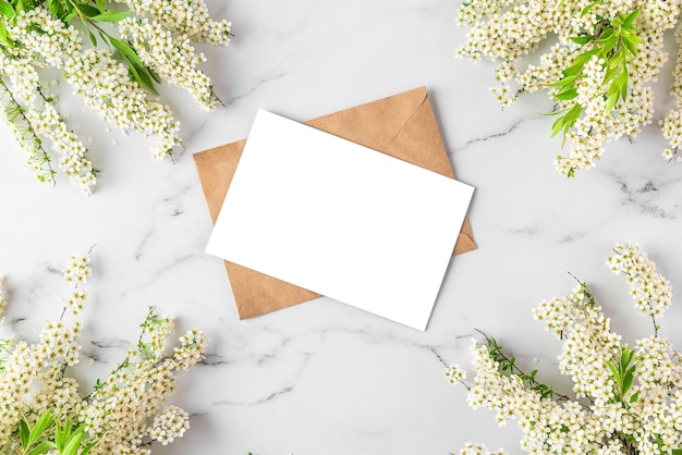 Blank greeting card in frame made of white flowers branches on white marble background mock up top view