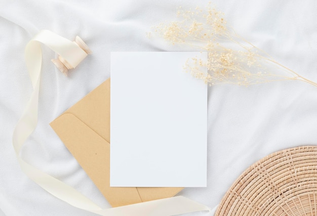 Blank greeting card invitation Mockup 5x7 on envelope with dry flowers and ribbon on white fabric background flat lay mockup