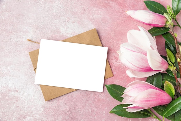 Blank greeting card with pink magnolia flowers and green leaves on pink concrete background Flat lay Mock up