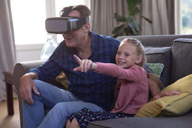 Blank meisje en haar grootvader genieten samen van hun tijd in een huis, zittend op een bank, senior man draagt een vr-headset, een meisje wijst in de lucht