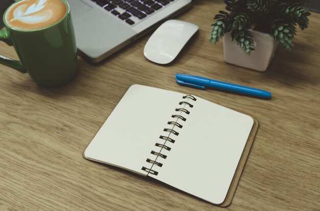 Blank notebook lying on a wooden table and a green coffee mug on the computer