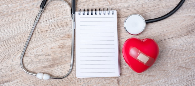 Taccuino in bianco, stetoscopio con forma rossa del cuore su fondo di legno.