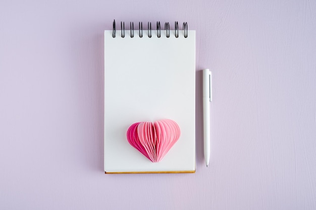 Пустой блокнот с ручкой и бумажным сердцем на сиреневом фоне День святого Валентина и День матери Копия