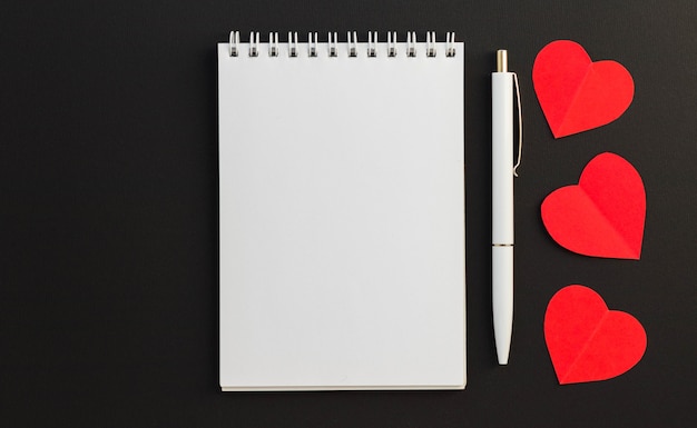 Blank notepad, pen and three red paper hearts on black background. Love message. Valentine's Day and romantic holiday concept. Top view, flat lay with copy space.