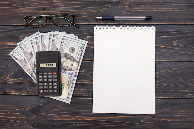 Blank notepad with pen, calculator and dollars on a wooden surface.