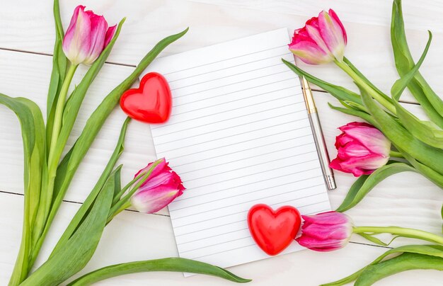 Blank notepad with two hearts and bouquet with tulips on the white wooden table Top view