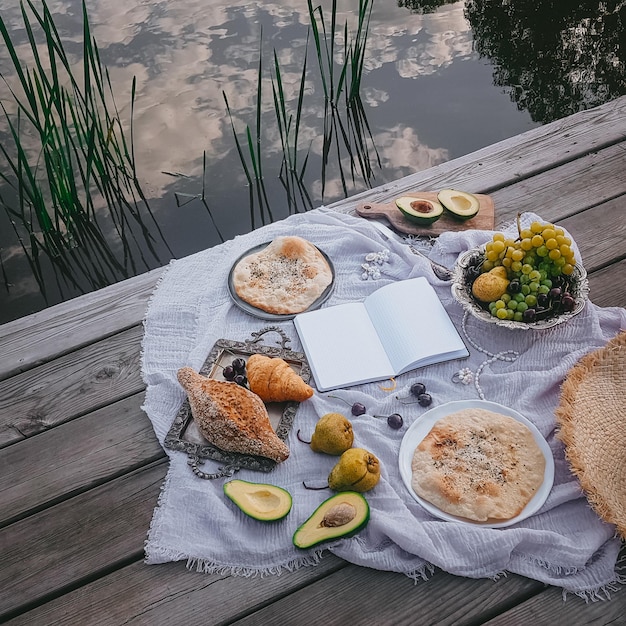 Blank notepad to write, Picnic in vintage style, Fruits and bread with vintage dishes, decorations on a wooden background near the water