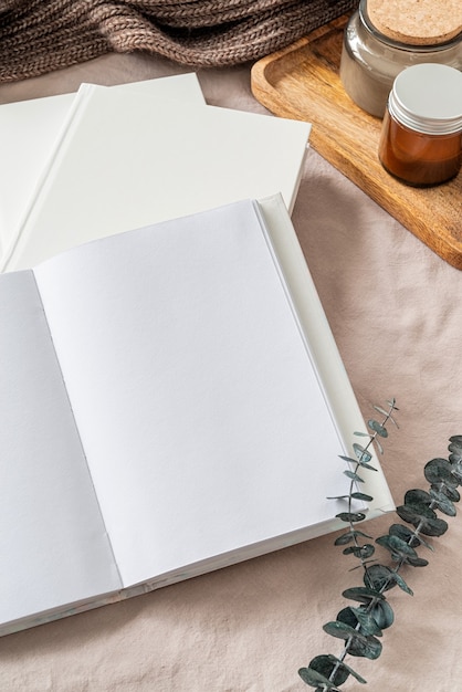 Blank opened book, candles and eucalyptus leaves on white bed, flat lay