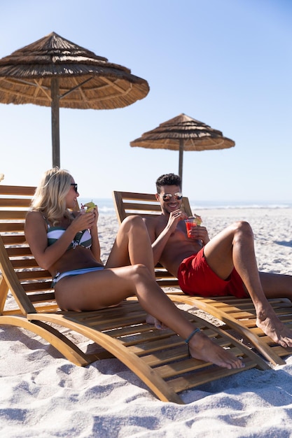 Blank paar genieten van tijd op het strand op een zonnige dag, zittend op ligstoelen en drankjes drinken met zee op de achtergrond