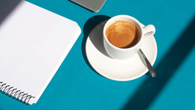 Blank page of a notebook next to a laptop and a white cup with coffee on a blue background