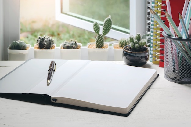 Blank page of notebook and pen on wooden table near open window with small cactus.