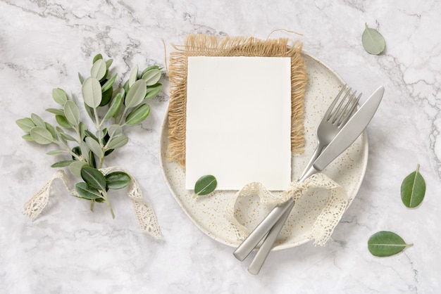 Blank paper card laying on white plate with fork and knife on marble table with eucalyptus branches and vintage ribbons around, top view. Card mockup