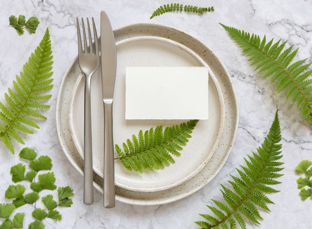 Blank paper card on table setting decorated with fern leaves on white marble table top view. Tropical mock-up scene with place card flat lay