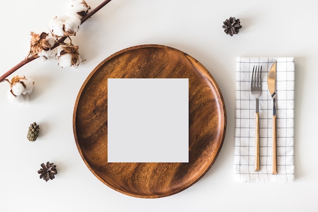 Photo blank paper greeting card on wooden dish, cutlery with checkered napkin, cotton flowers