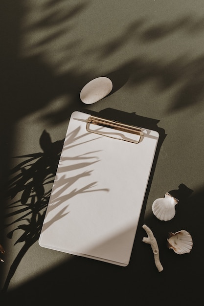 Фото Пустой лист бумаги с буфером обмена, цветочные тени солнечного света, камень, морская ракушка, коралл на темно-зеленом