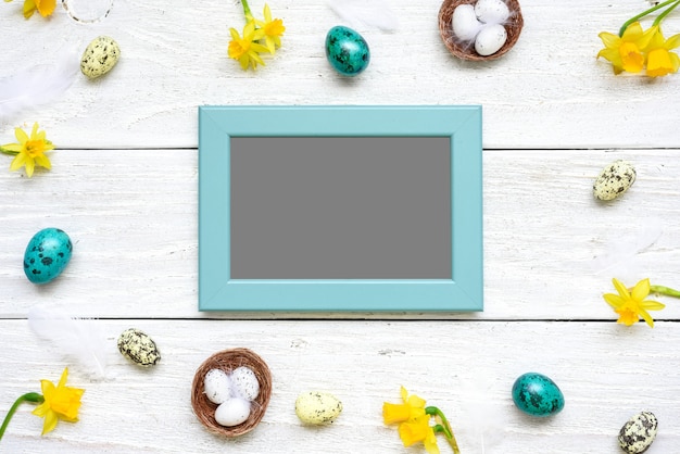 Blank photo card in frame made of quail eggs, spring flowers and feathers on white wooden table