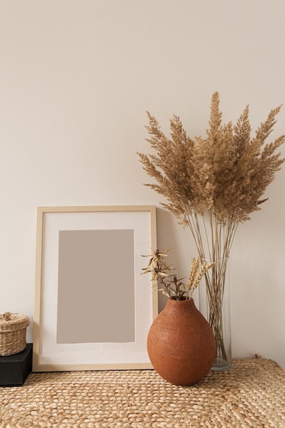 Blank photo frame, reeds bouquet in vase, rye in clay pot at white wall