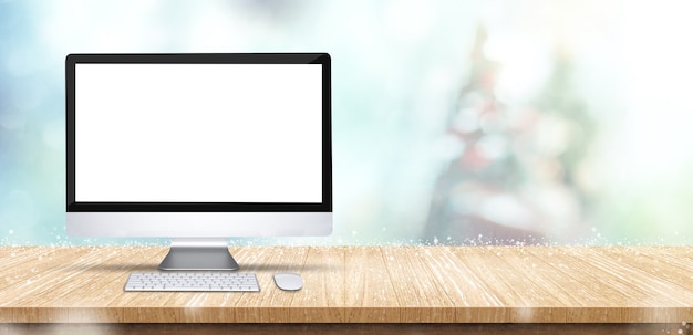 Blank screen computer desktop on wooden table with abstract blur christmas tree and snow falling background with bokeh