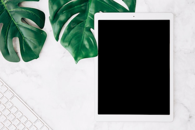 Blank screen digital tablet with monstera leaves and keyboard on white marble desk