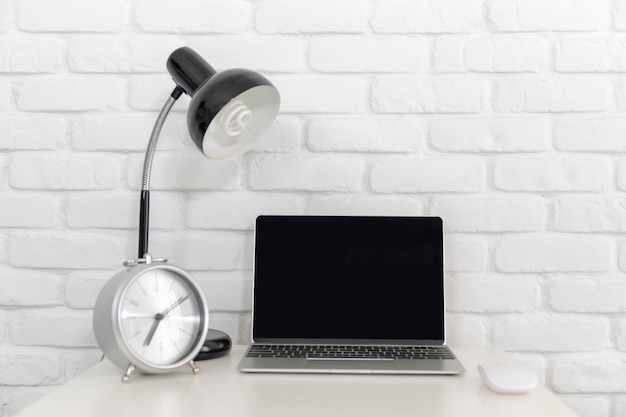 A blank screen Laptop computer on white desk with lamp and white brick wall.