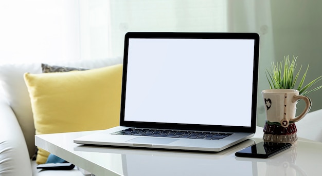 Blank screen laptop with smartphone, mug and houseplant on white wooden top table in living room.