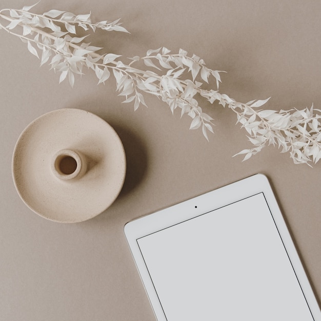 Blank screen tablet, white floral branch on neutral pastel beige background. Flat lay, top view.