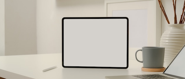 Blank screen tablet on white worktable with stylus pencil, laptop, coffee cup and decoration