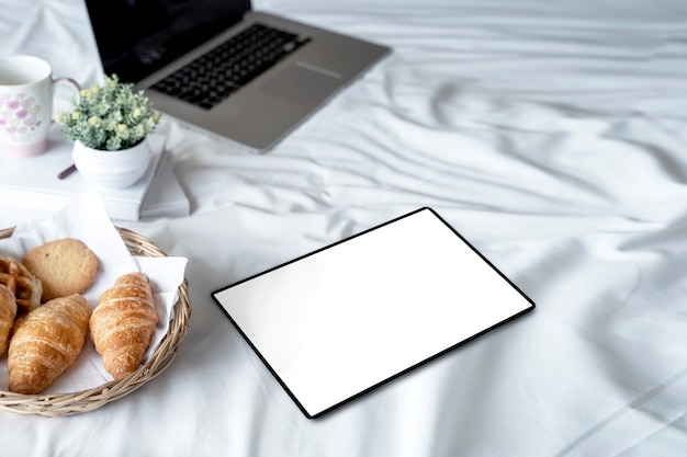 Blank screen tablet with croissant bread on white bed sheets.
