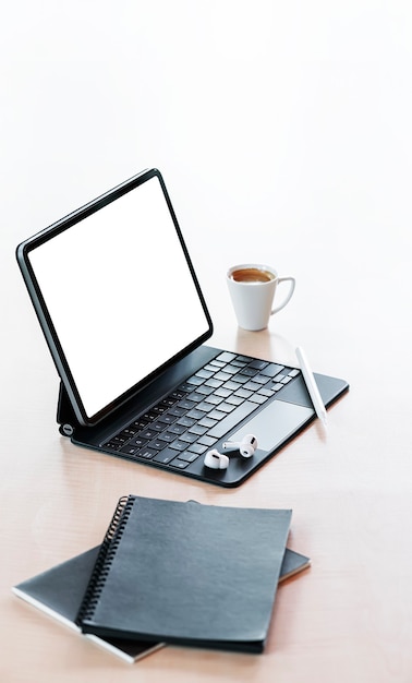 Tablet schermo vuoto con tastiera magica, tazza di caffè e notebook su sfondo bianco, vista verticale.