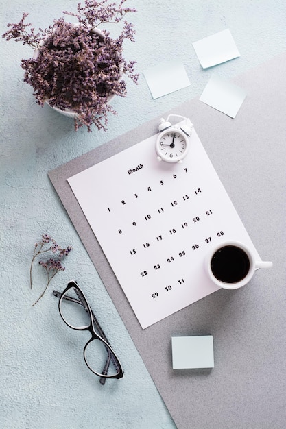 Blank sheet of monthly calendar, glasses, coffee cup and alarm clock on the desktop. Time planning and organization. Vertical view