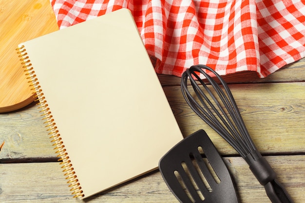 Blank sheet of opened notepad and kitchen utensils on  table with tablecloth, copy space