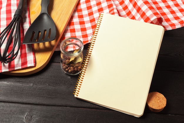 Blank sheet of opened notepad and kitchen utensils on  table with tablecloth, copy space