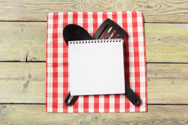 Blank sheet of opened notepad and kitchen utensils on  table with tablecloth