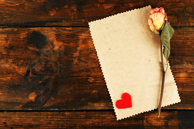 Photo blank sheet of paper with little hear and dried rose on wooden table background