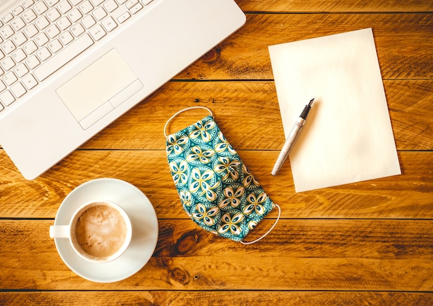 A blank sheet of paper with pen on a wooden work table with a coffee cup and protective face mask.