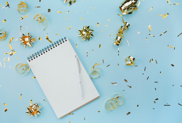 Blank spiral notebook with white pen on a blue background with golden confetti and bows. Fashionable color and style. Flat lay. Copy space. Blogger idea.