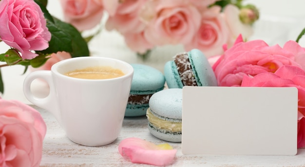 Blank white business card and cup with espresso coffee and white ceramic cup with coffee and blue macaron on a white table, behind a bouquet of pink roses