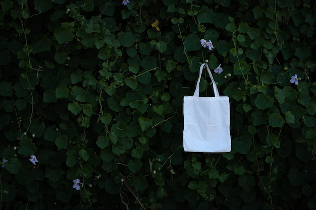 Пустая белая тканевая сумка-тоут на фоне листвы деревьев зеленого куста